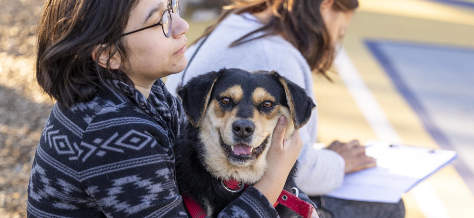woman wearing patterned sweater sits outside with a black and tan dog in her lap